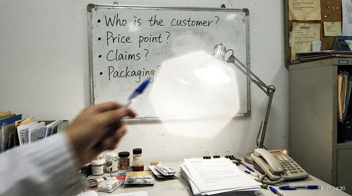 A person writing on a whiteboard with questions like 'Who?', 'What Price?', 'Claims?'.