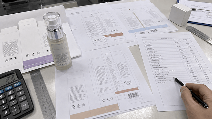Label artwork proofs and a printed ingredient list spread across a desk for market-by-market review, iPhone-angle snapshot