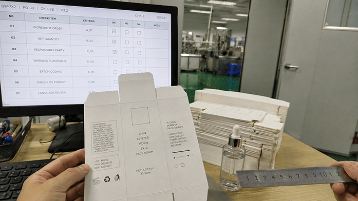 A printed label proof held up against a screen showing ingredient and net quantity requirements, iPhone-angle snapshot